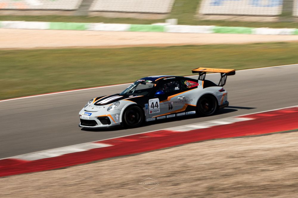 White Porsche race car number 44 with orange spoiler speeding on a professional racing circuit track.