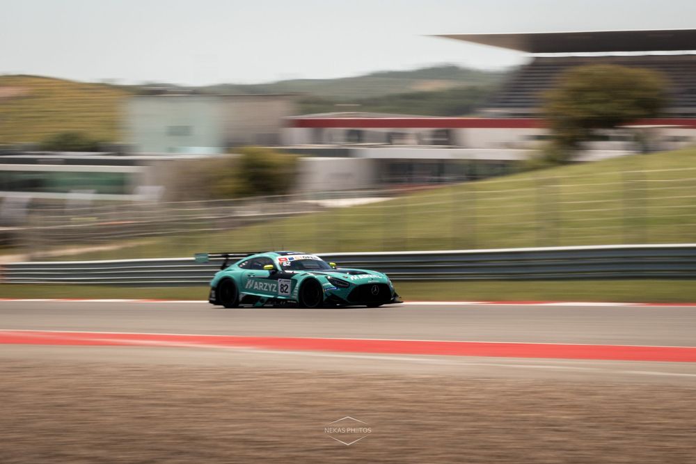 Teal racing car speeding on a professional circuit track with red pit lane buildings in the background.