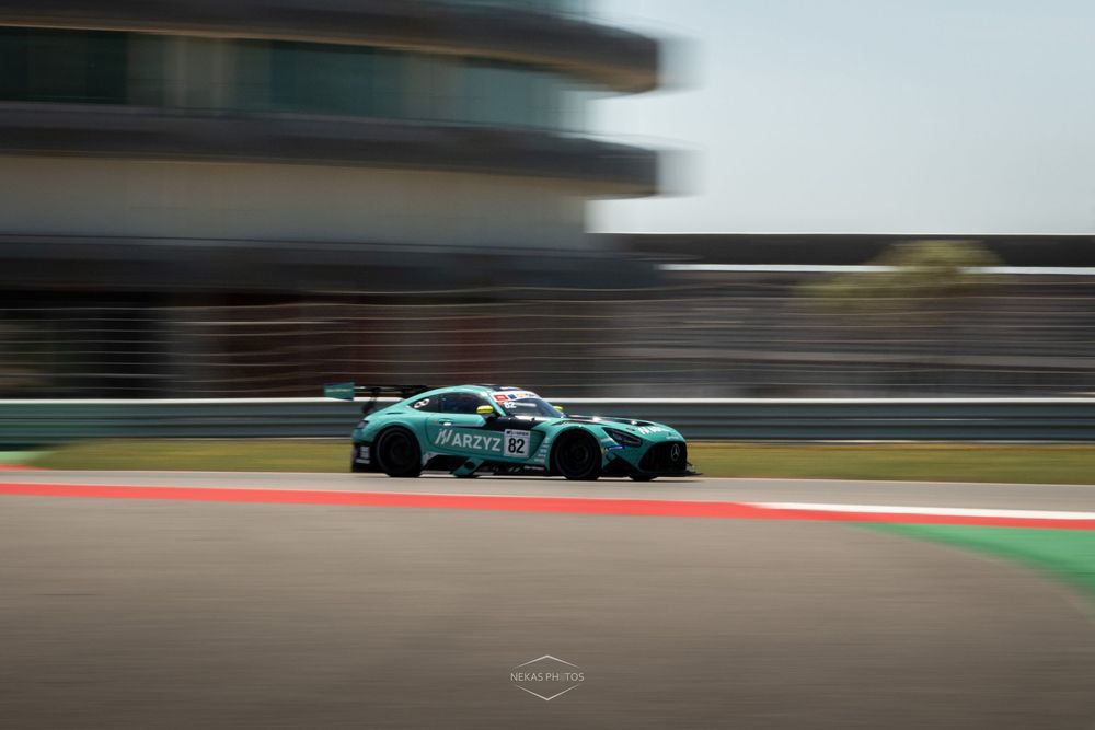 A teal racing car speeds around a professional motorsport circuit, captured with motion blur emphasizing its high velocity.