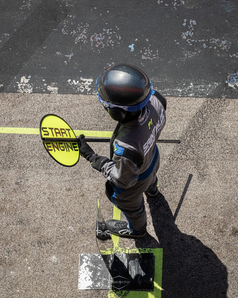 Overhead view of a racing pit crew member in helmet holding a yellow START ENGINE sign on a race track.