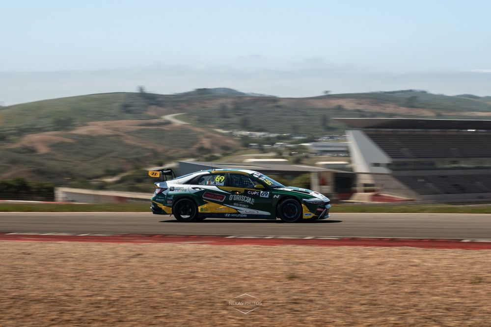 A black and yellow racing car speeding on a circuit track with hills and grandstands in the background.