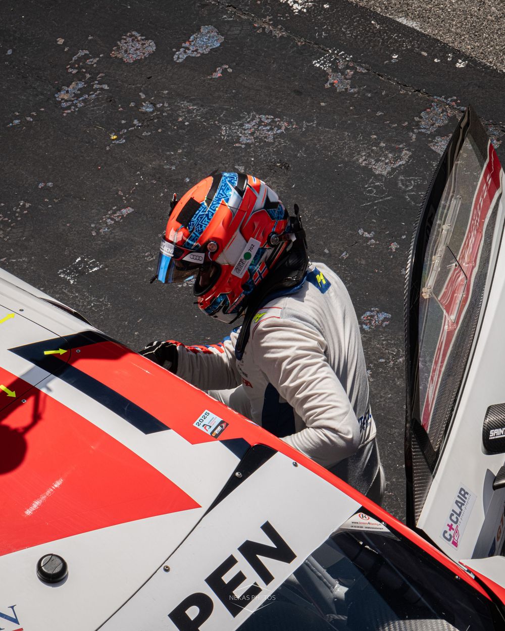 Racing driver in orange helmet climbs into a red and white race car cockpit on a motorsport circuit.