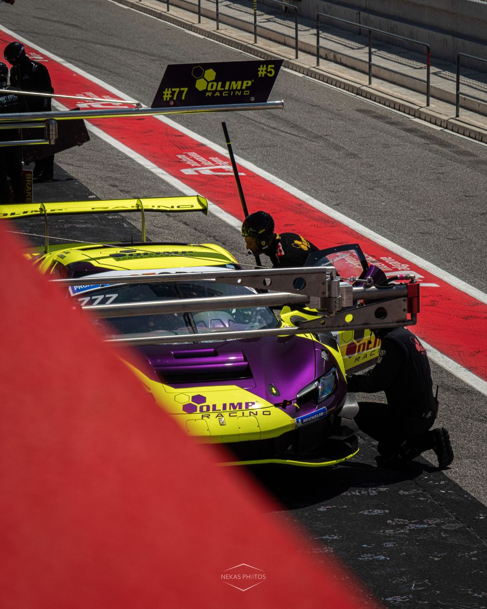 Olimp Racing pit crew services a yellow and purple race car #77 in the pit lane during a motorsport event.