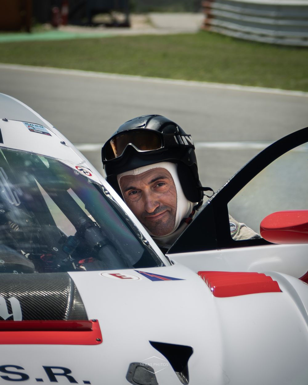 Race car driver in white helmet and goggles smiling from cockpit of white and red racing car on track.