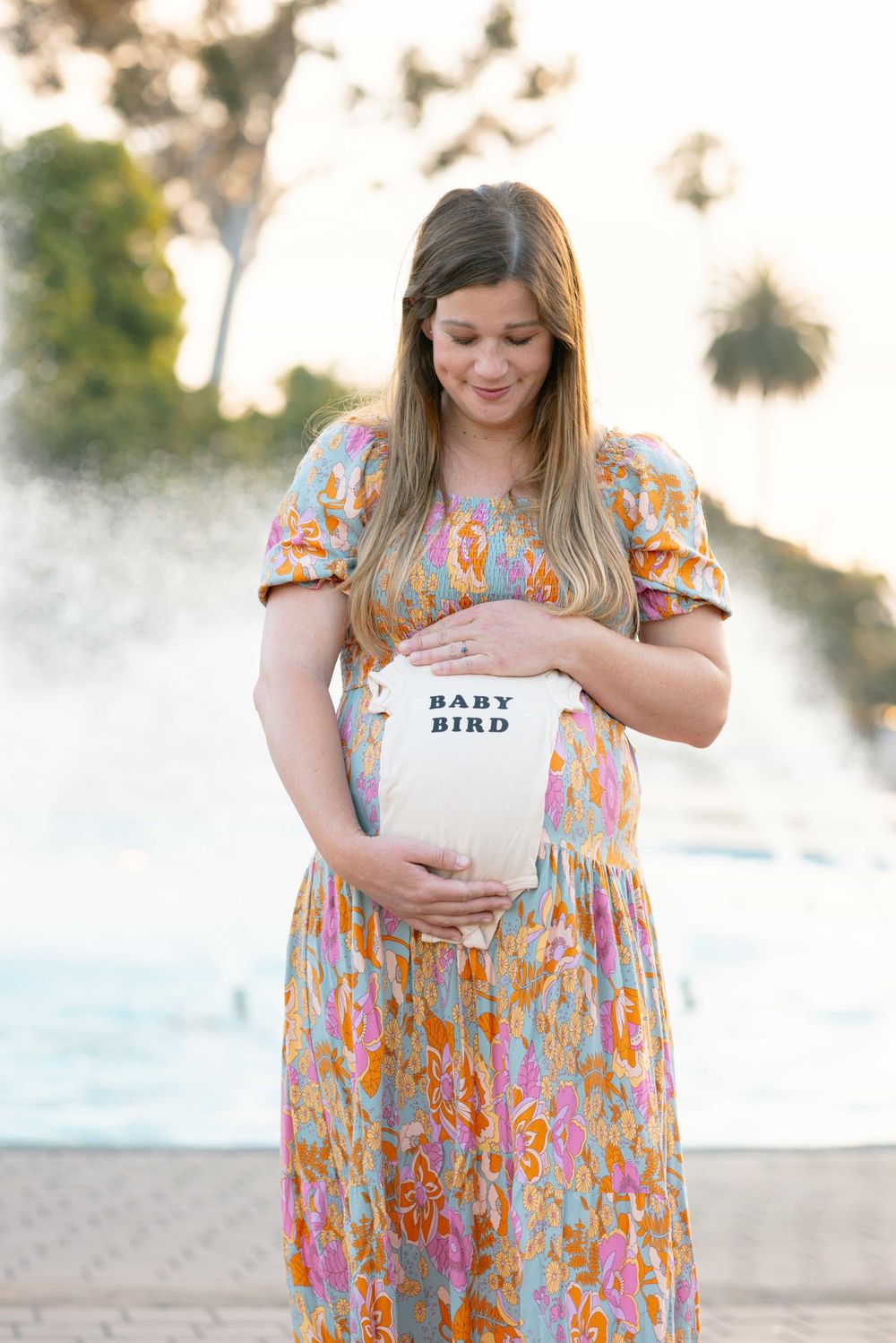 Pregnant woman in colorful floral dress tenderly holds baby bird onesie against her baby bump near fountain.