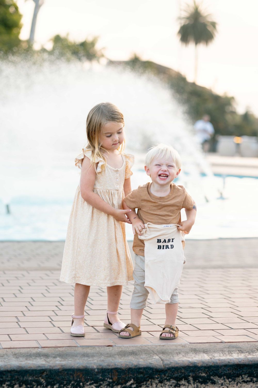 Two young siblings stand by a fountain, girl in yellow dress holds boys hand as he laughs holding baby onesie.
