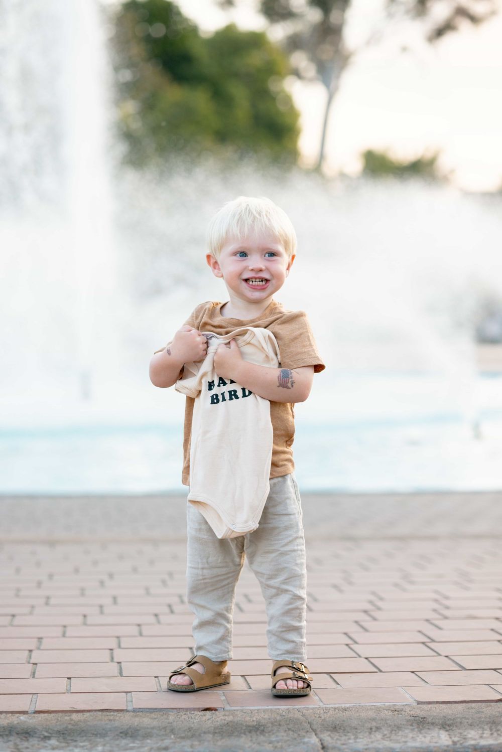 Cheerful blonde toddler boy holds baby bird onesie while standing in front of a sparkling fountain outdoors.