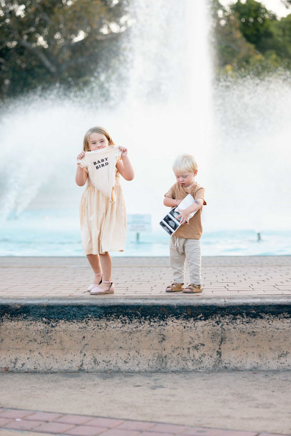 Two young children stand by fountain, girl holds baby bird onesie while boy examines ultrasound photo.