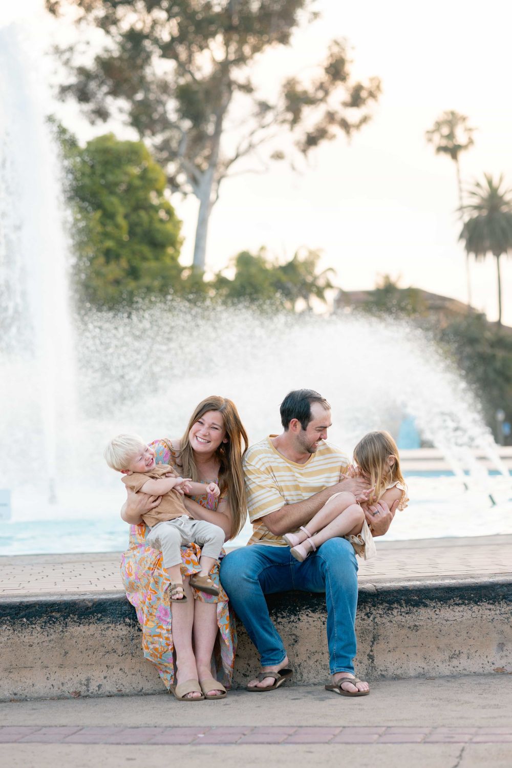 Happy family of four sits together by a fountain, pregnant mother and father holding their two young children.