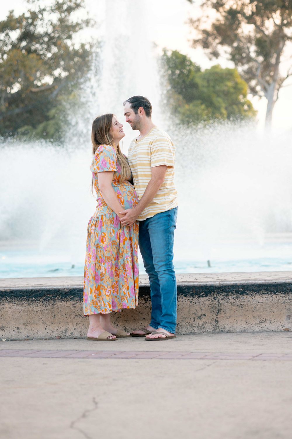 Expecting couple shares a romantic moment gazing at each other in front of a large outdoor fountain.