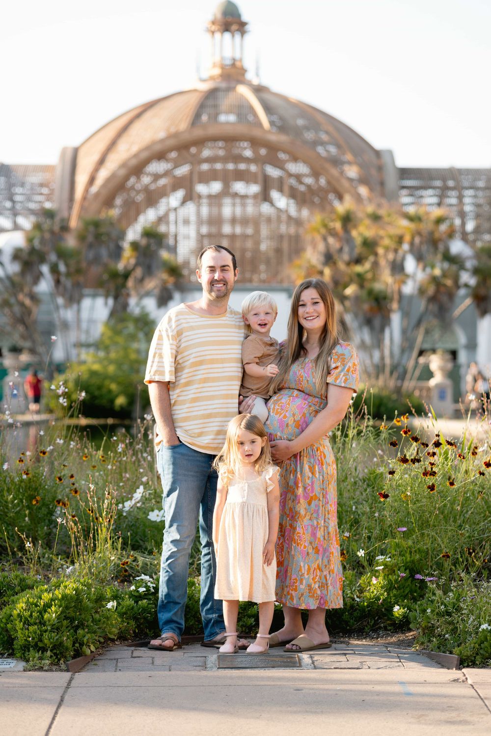 Joyful family of four poses in garden with pregnant mother, father holding toddler, and young daughter smiling.