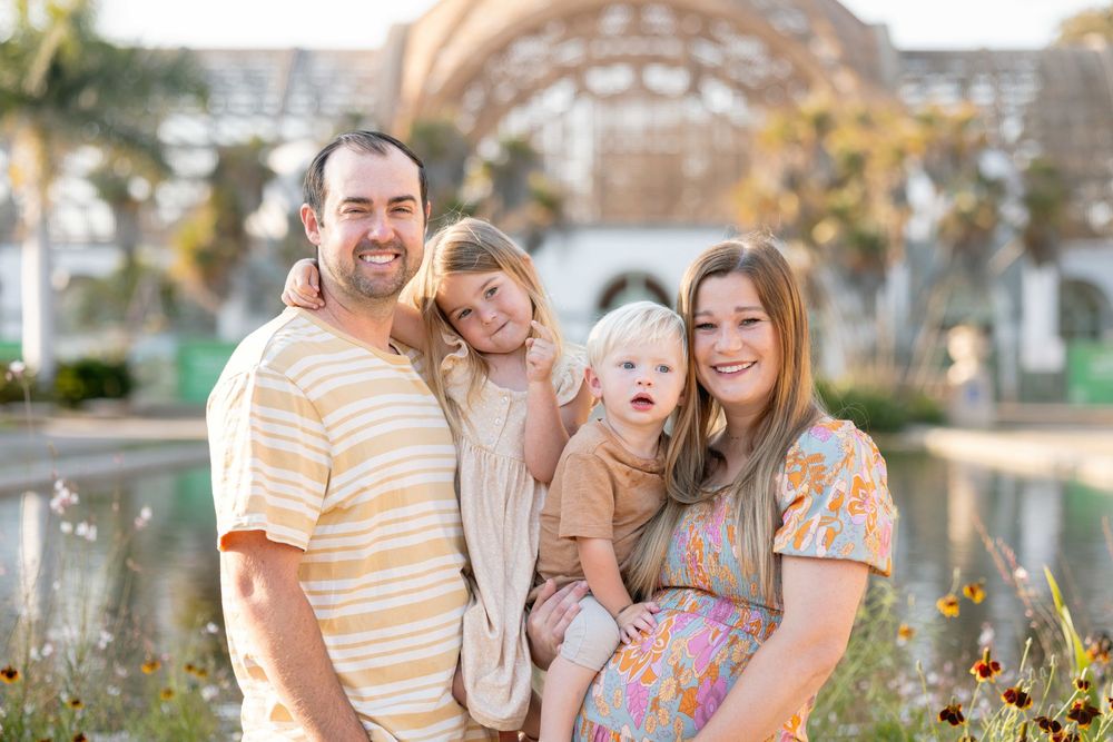 Happy family of four posing outdoors near a garden, parents smiling with two young children in warm golden light.