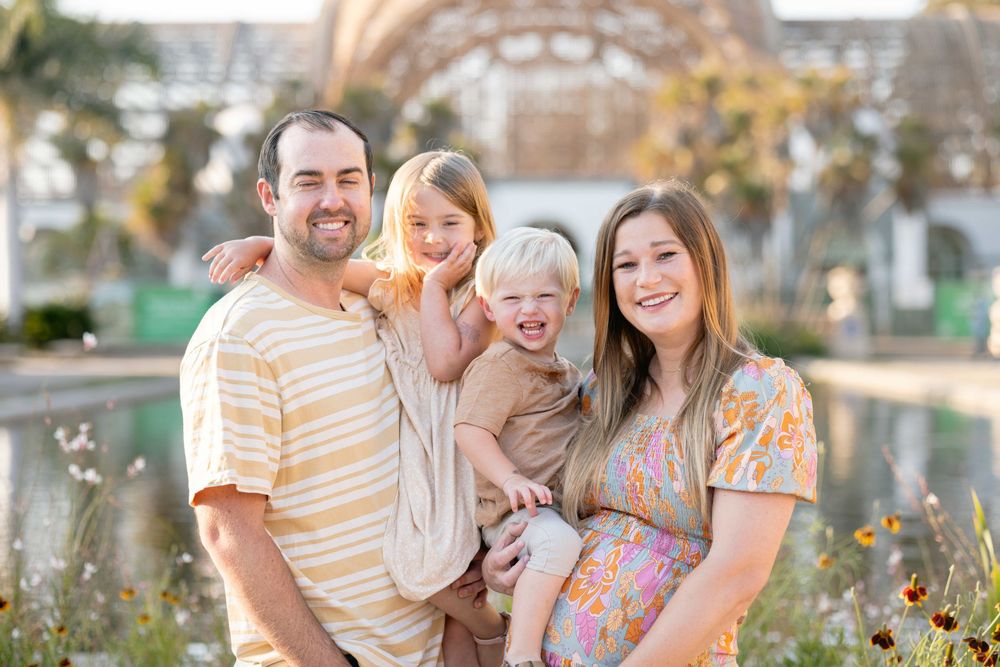 Joyful family portrait with parents holding two laughing children in a sunny outdoor garden setting.