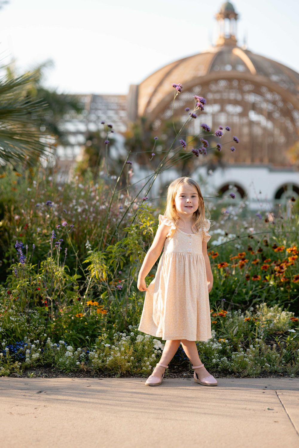 Adorable blonde girl in a peach dress standing in a colorful garden with a domed building behind her.