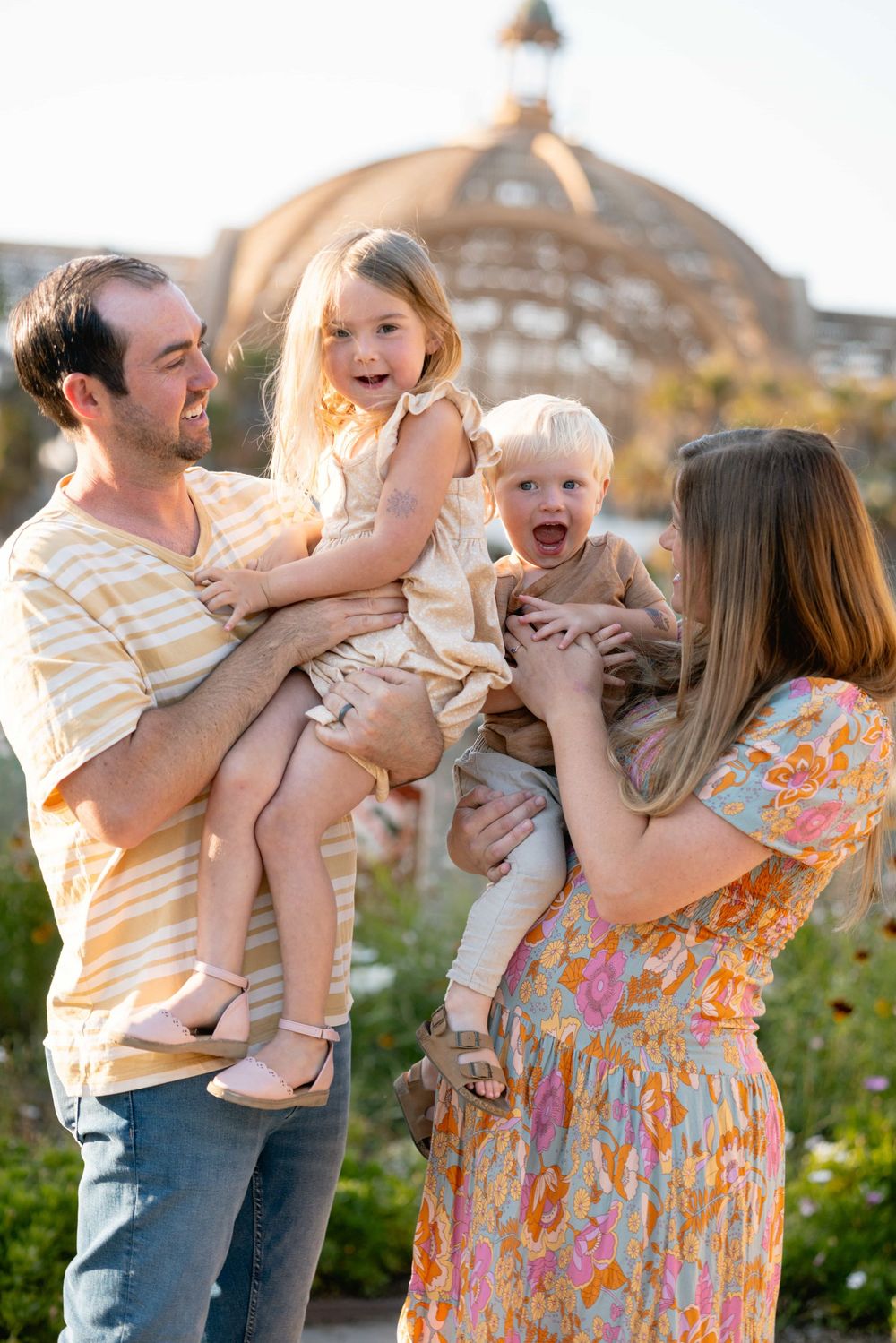 Parents each holding a laughing child outdoors with a large domed building visible in the background.