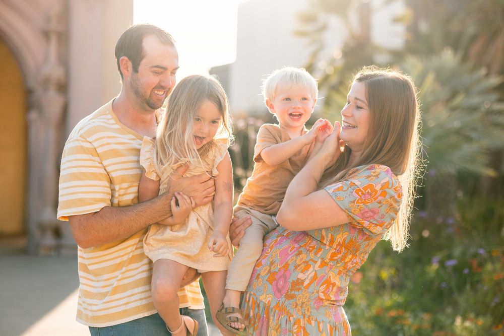 Smiling family of four in golden sunlight, parents holding two young children in an outdoor setting.