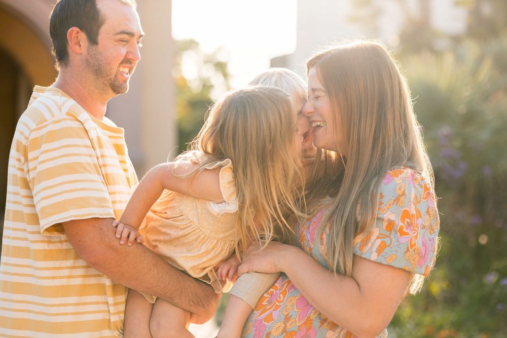 Tender moment of a mother hugging her young daughter while father smiles in warm sunlit outdoors.