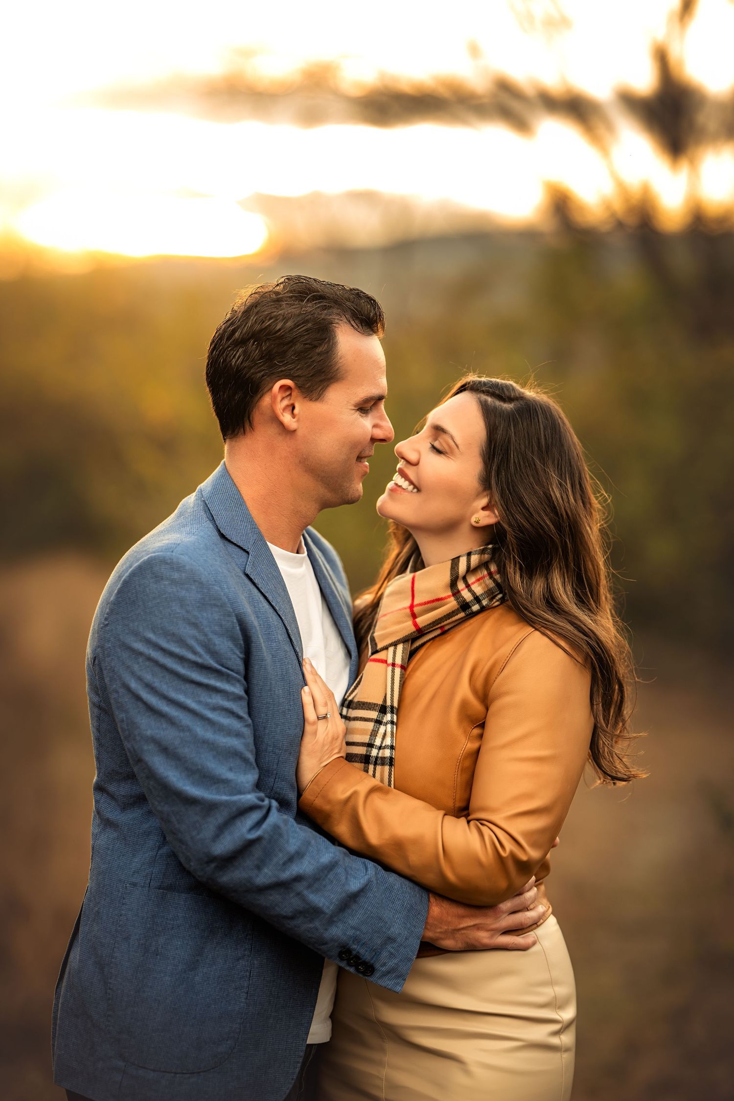 Couple embracing and smiling at each other at sunset in a golden autumn outdoor setting, woman in Burberry scarf and man in blue blazer.