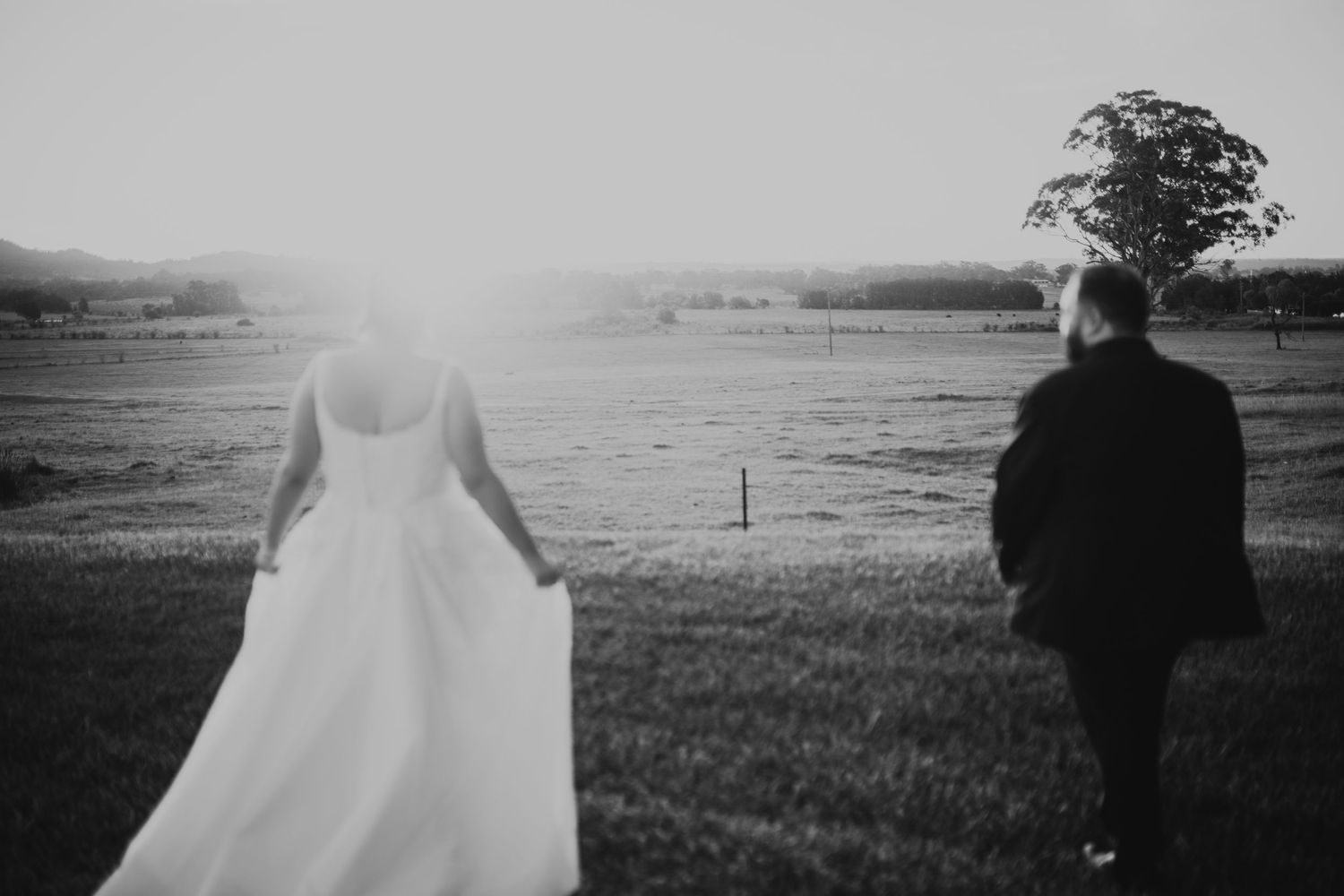 Black and white photo of a bride and groom walking hand in hand through a sunlit field on their wedding day.