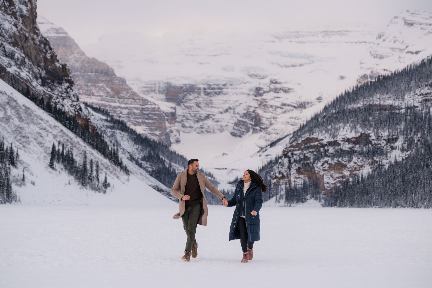 Couple holding hands on snowy frozen lake with majestic mountain backdrop in winter at Lake Louise, Banff.