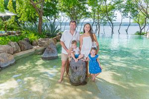 Family of four posing by a tropical pool with lush greenery and calm water in the background.