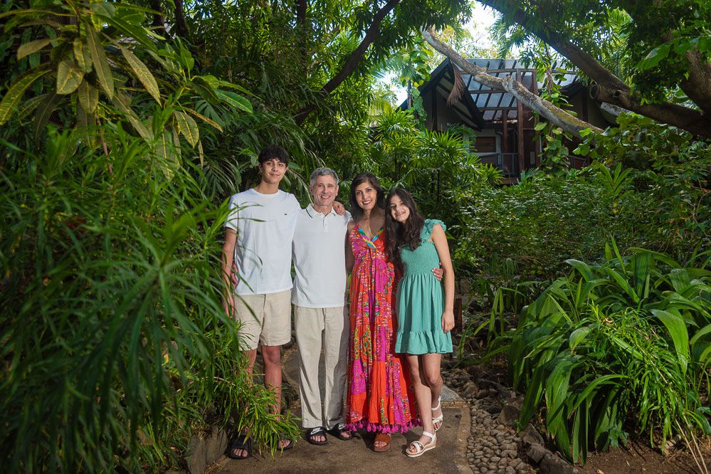 Family of four posing on a garden path surrounded by lush tropical greenery near a wooden cottage.
