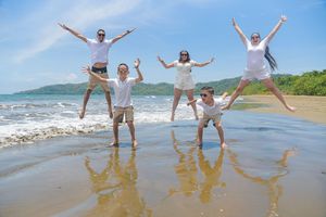 Family of five jumping joyfully on a tropical beach with blue skies, waves, and lush green hills in the background.