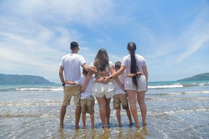 Family of five in white outfits standing together on a tropical beach shore, viewed from behind with ocean waves.