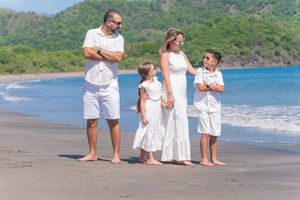 Family of four in matching white outfits posing on a tropical beach with lush green hills and blue ocean.