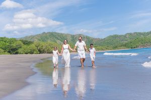 Family of four in white outfits walking on a tropical beach with lush green hills and blue sky.