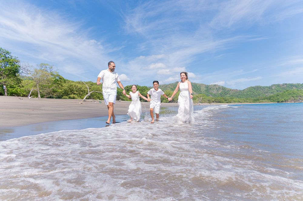 Family in white outfits running joyfully along a tropical beach with lush green hills and blue sky.