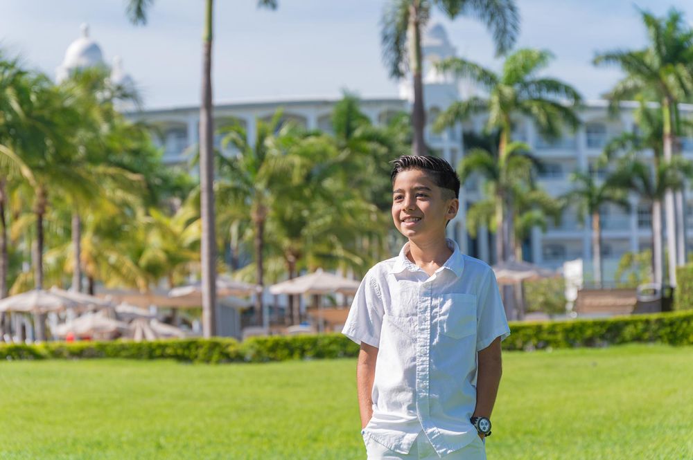 Young man smiling in white outfit on green lawn with palm trees and resort building in background.