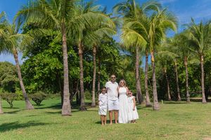 Family of four in white outfits posing on green lawn surrounded by tall palm trees under a bright blue sky.
