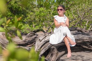 Young girl in a white dress and sunglasses sitting on driftwood surrounded by lush green trees.