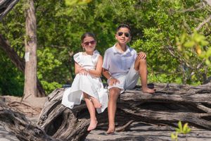 Two children in white outfits and sunglasses sit on driftwood logs surrounded by lush green trees on a tropical beach.