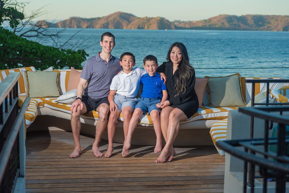 Family of four sitting together on an outdoor deck with a scenic ocean and mountain view at sunset.