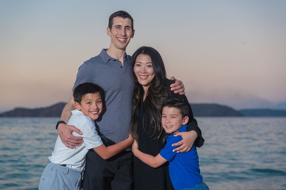 Family of four smiling and hugging at a beach during sunset with ocean and islands in the background.