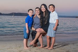 Family of four smiling together on a sandy beach at sunset, with calm ocean waters and a sailboat in the background.