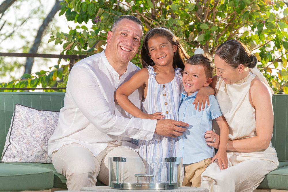 Happy family of four smiling together on outdoor patio sofa surrounded by lush green trees.