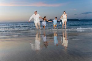 Family of four running joyfully on a beach at sunset, reflecting on wet sand with ocean waves in the background.