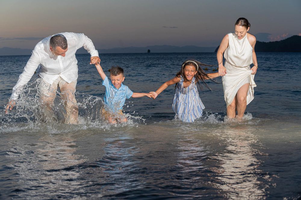 Family of four splashing joyfully in ocean waves at sunset on a tropical beach.