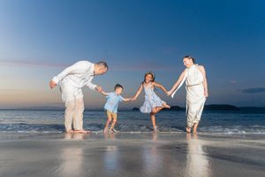 Family of four holding hands and playing on a sandy beach at sunset with ocean waves in the background.