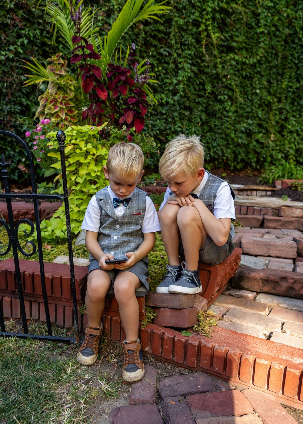 Two young boys in matching plaid vests sit on brick steps in a garden, looking at a smartphone together.