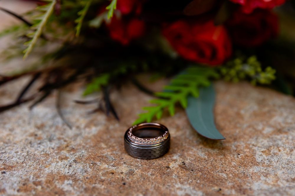 Dark tungsten wedding band with rose gold detail rests on stone surface beside red rose bridal bouquet.