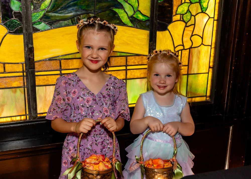 Two young flower girls in dresses holding baskets of flowers pose before a colorful stained glass window.