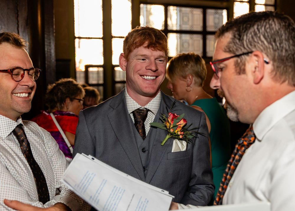 Three men in formal attire smile and converse at a wedding reception indoors.