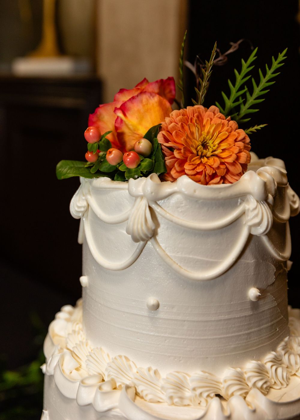 Elegant white wedding cake topped with orange dahlia, pink roses, and greenery, featuring draped fondant details.