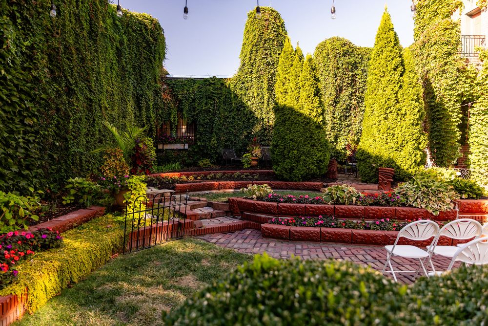 Lush terraced garden with ivy-covered walls, colorful flowers, and white chairs in warm sunlight.