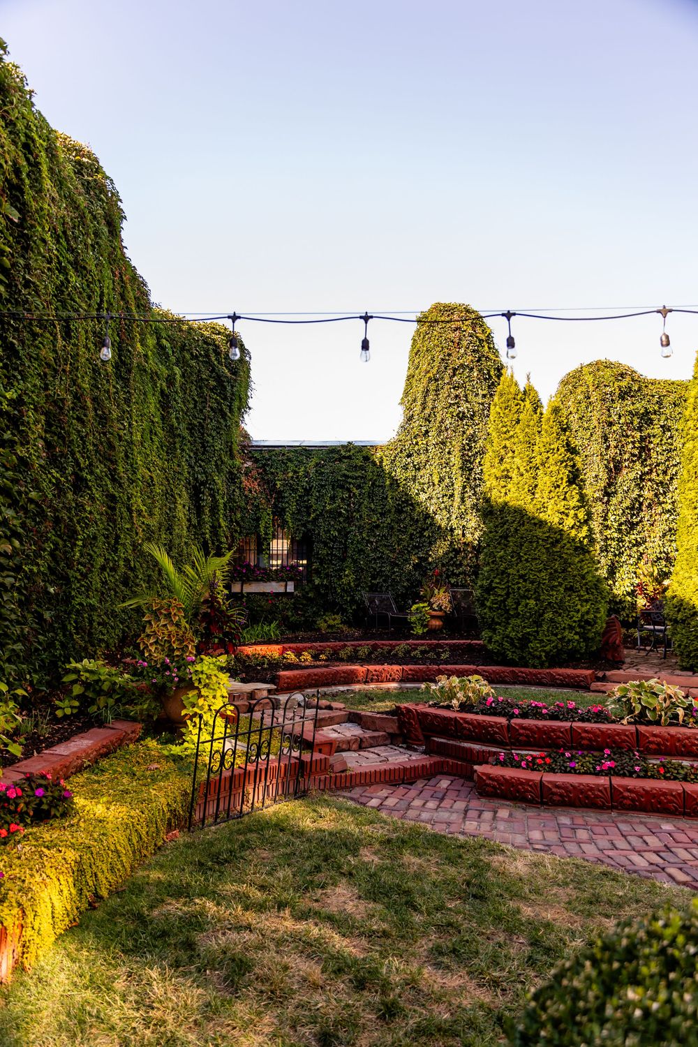 Lush garden with ivy-covered walls, manicured hedges, brick pathways, colorful flowers, and string lights under a clear sky.