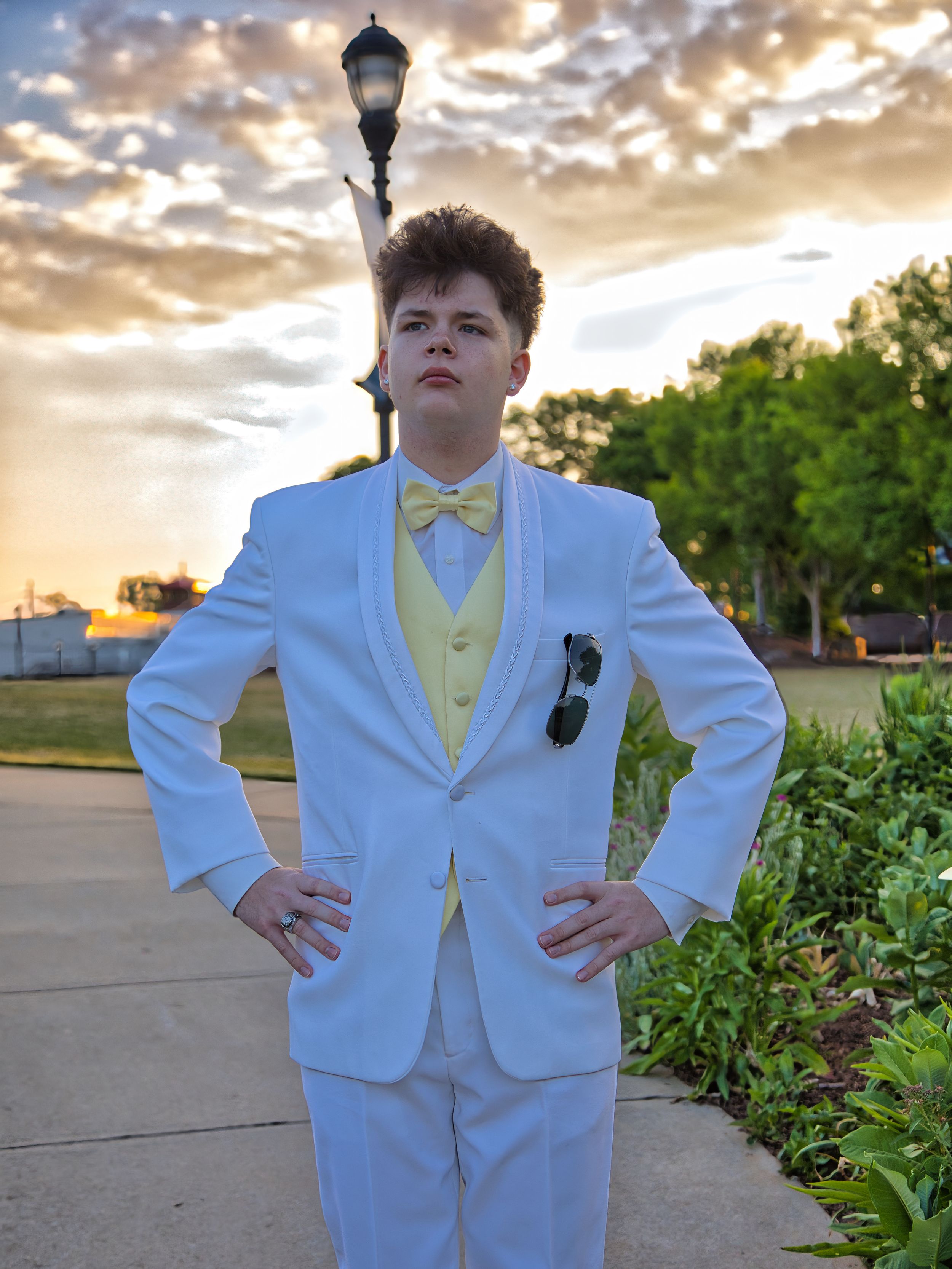 Young man in white tuxedo with yellow bow tie posing outdoors at sunset for prom night.